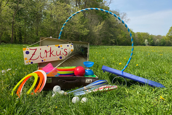tereza-kreim-092 Auf einer Frühlingswiese steht ein Koffer an dem ein buntes Schild hängt, auf diesem steht "Zirkus". Im Koffer und drum herum stehen und liegen Zirkusrequisiten: Keulen, Jonglierteller, Jongliertücher, Bälle, Ringe, ein Hula Hoop, ein Diabolo und ein Rola Bola.